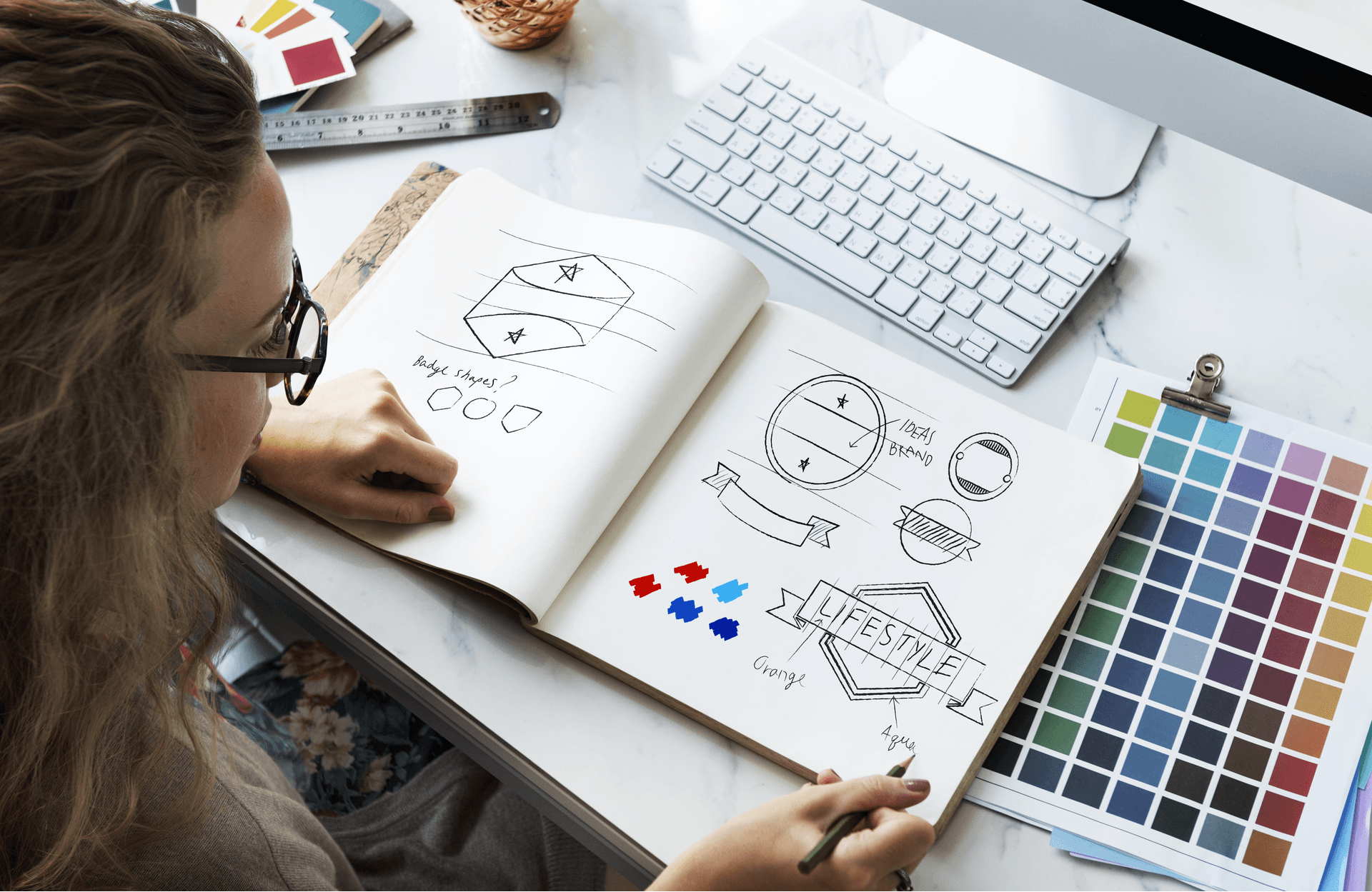 person drawing logos in a sketchbook next to a color palette