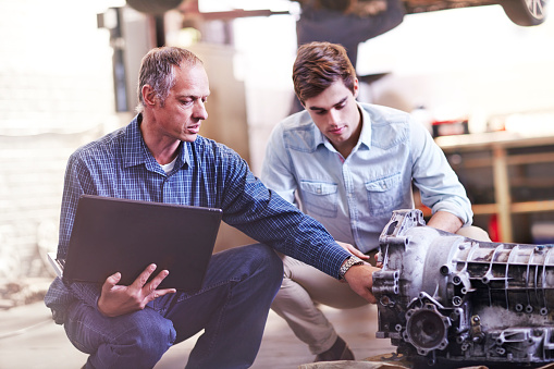 Photo of two men working over an auto engine. One man holding laptop.